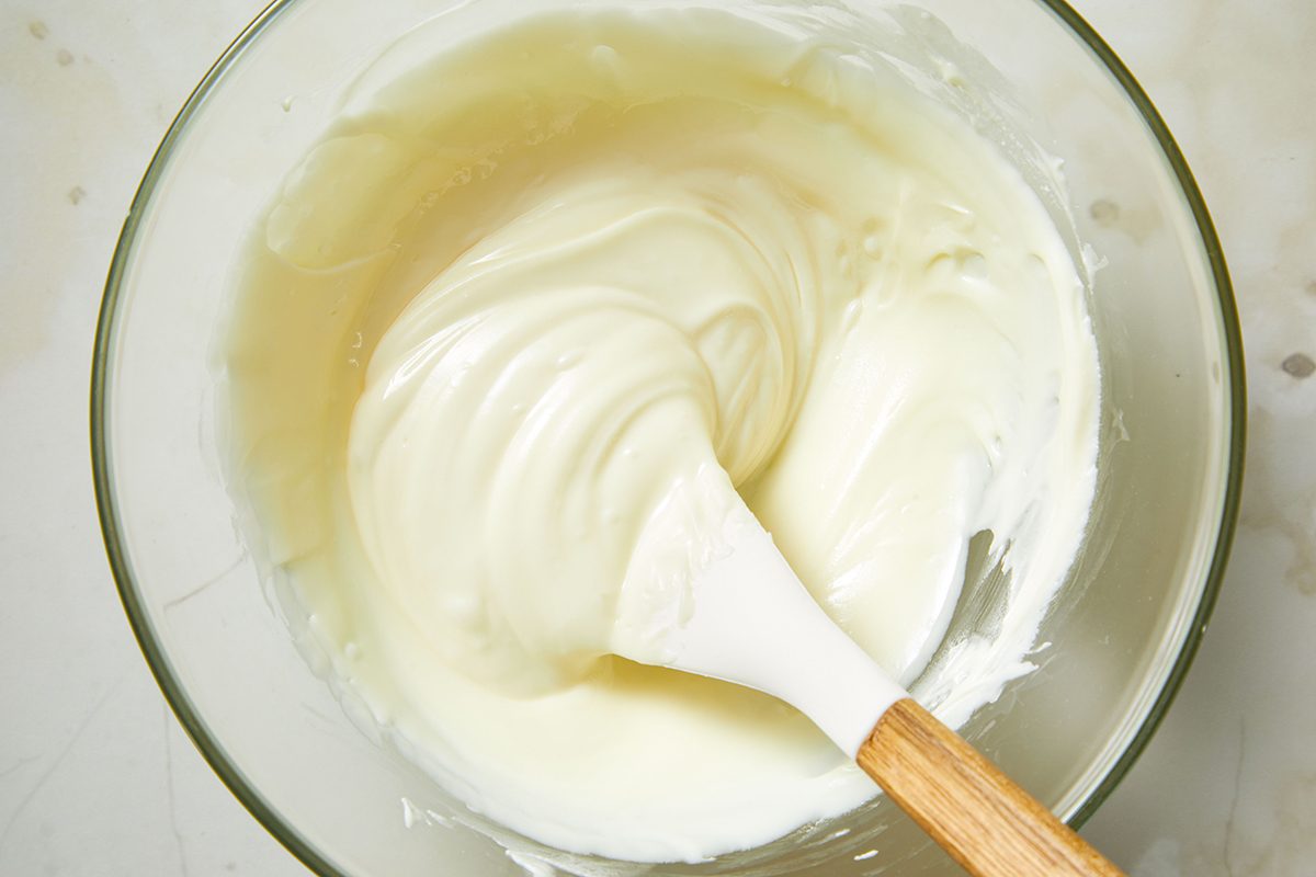 A glass bowl filled with creamy, smooth white mixture being stirred with a white spatula that has a wooden handle.
