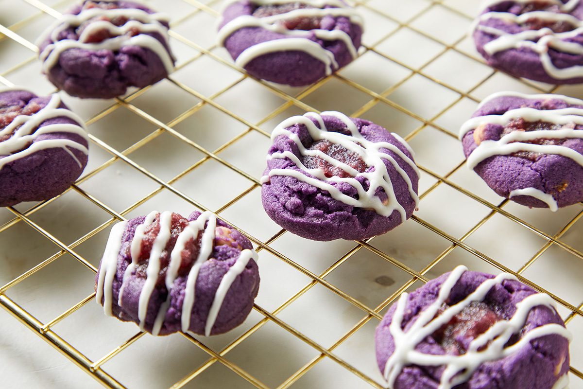 Eight vibrant purple cookies with white icing drizzle are cooling on a gold wire rack, placed on a white surface.