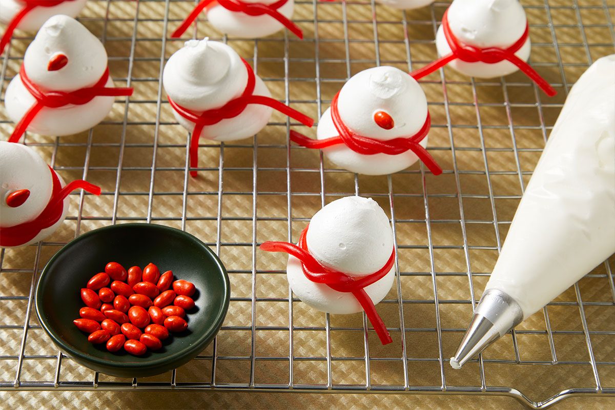 3/4th shot of Meringue snowmen with red licorice scarves and red candies cool on a wire rack A piping bag and bowl of red candies sit nearby