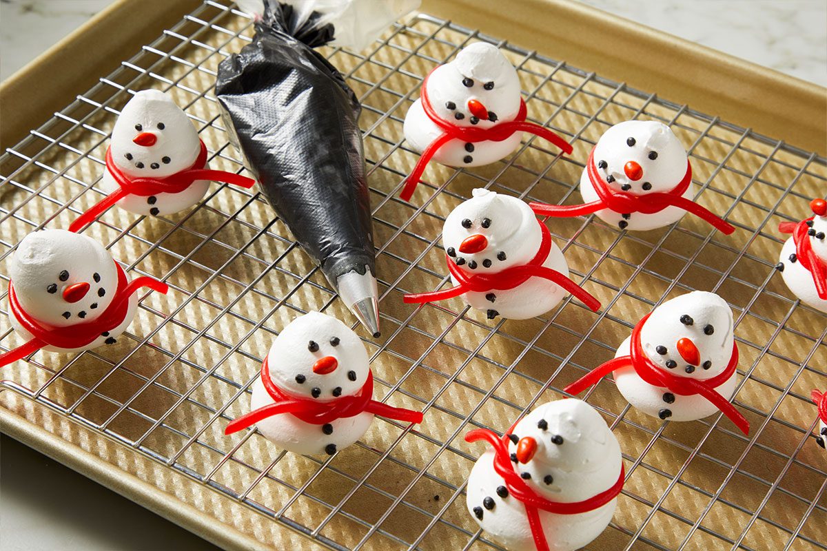 3/4th shot of 3D Meringue Snowmen are shaped like snowmen with red candy scarves and icing faces sitting on a wire rack over a baking sheet while a piping bag with a metal tip rests among them