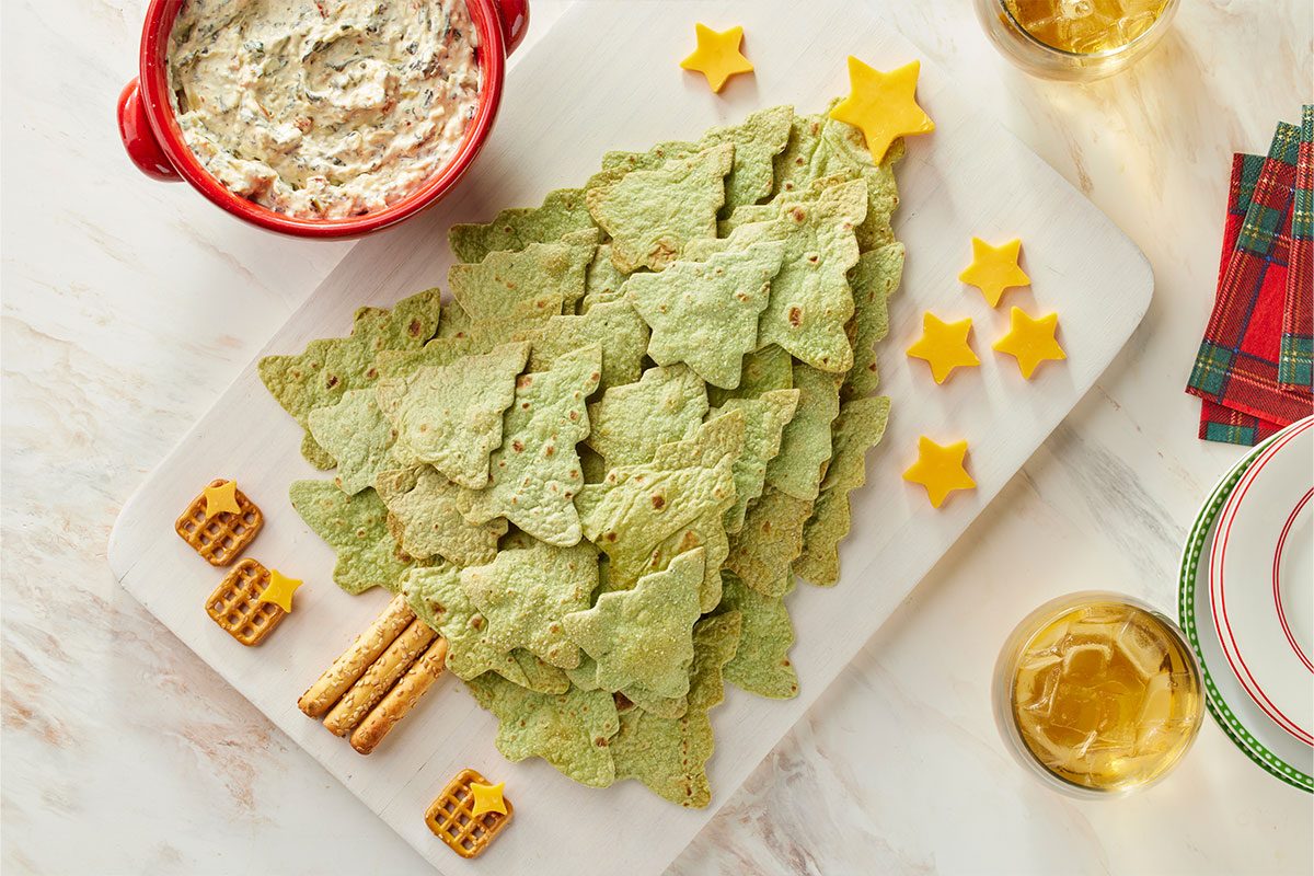 Overhead shot of Christmas tree shaped green tortilla chips arranged into a festive tree on a serving board, complete with pretzel-stick trunks and cheddar cheese stars, with a holiday dip and drinks nearby;