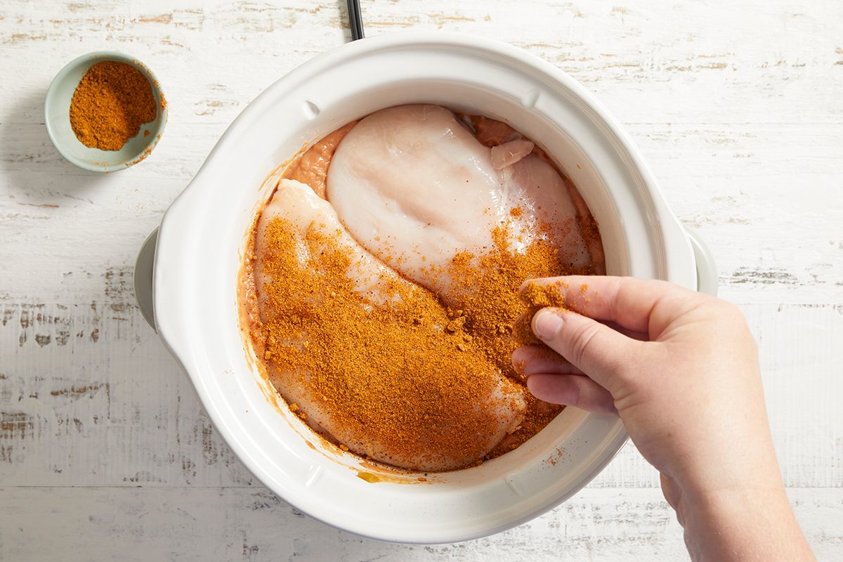 A hand sprinkles orange-colored seasoning onto raw chicken breasts in a white slow cooker. A small bowl of additional seasoning sits nearby on a white surface.