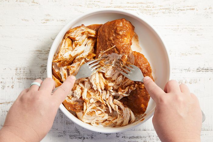 Two hands use forks to shred cooked, seasoned chicken breasts in a white bowl on a light wooden surface.