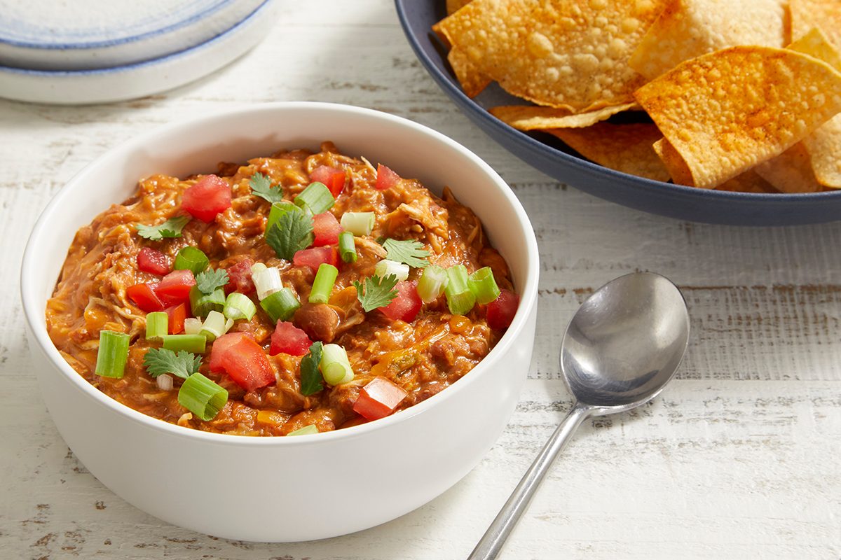 A white bowl of cheesy bean dip topped with chopped tomatoes, green onions, and cilantro, sits next to a plate of crispy tortilla chips and a spoon on a white wooden table.