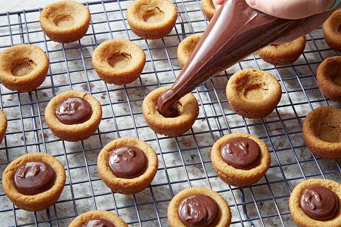 A hand uses a piping bag to fill cookie cups with chocolate filling on a wire cooling rack, with several cookie cups already filled and some still empty.