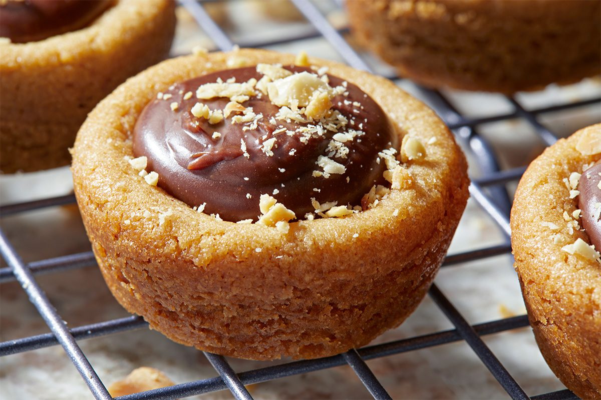 A close-up of a peanut butter cookie cup filled with chocolate and topped with crushed peanuts, resting on a metal cooling rack.