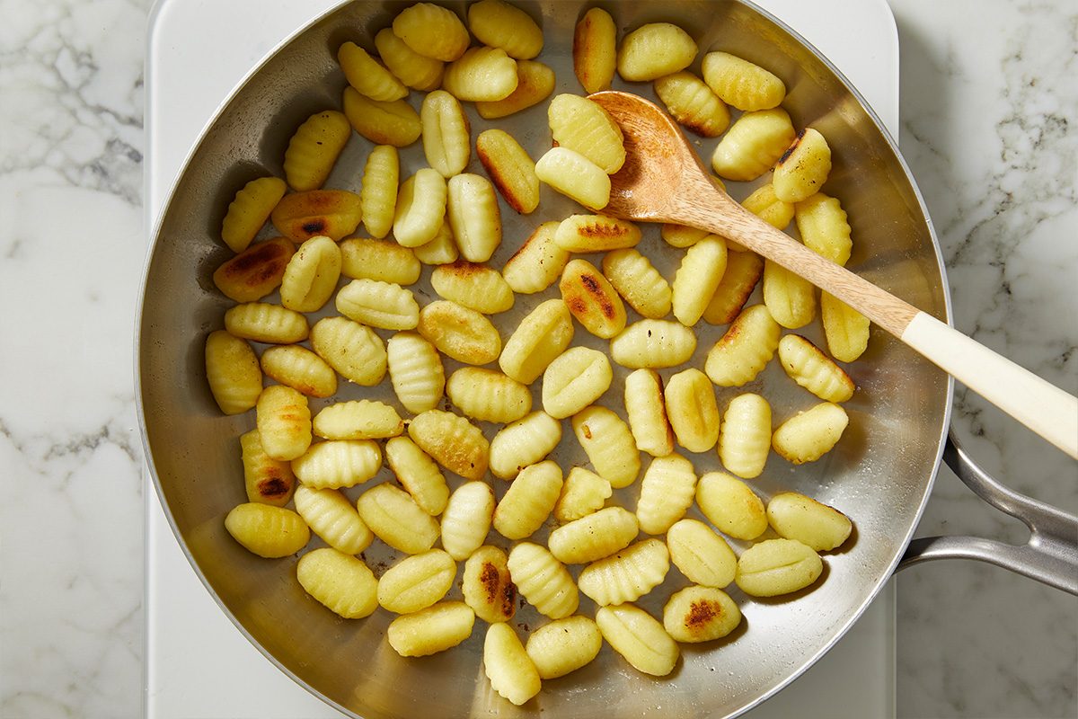 Overhead shot of golden gnocchi sautéing in a skillet, being stirred with a wooden spoon on a stovetop;