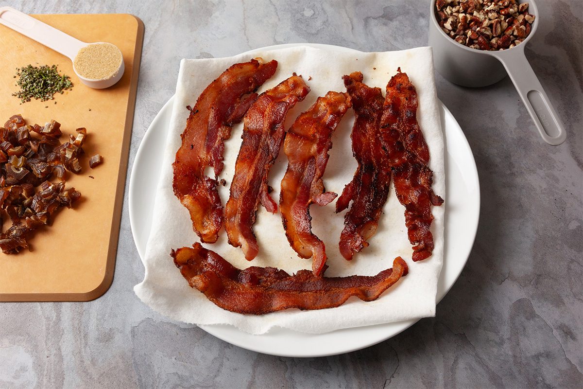 3/4th shot of a plate lined with paper towels holds seven cooked bacon strips and beside it are chopped dates seasoning and pecans on a gray stone surface