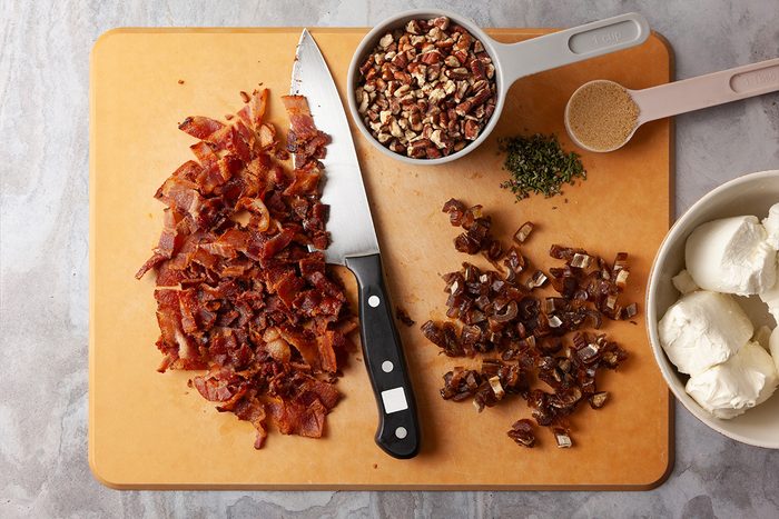 Overhead shot of a gray countertop a cutting board displays chopped bacon chopped dates and herbs with a knife Nearby are pecans brown sugar and cream cheese
