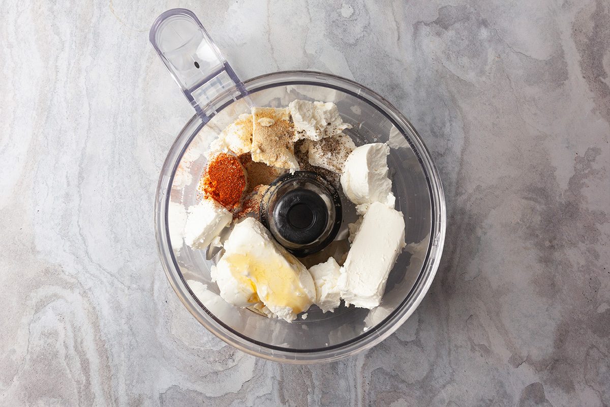 Overhead shot of a food processor bowl appears from above on a light marble surface you see cream cheese butter and a blend of colorful spices inside