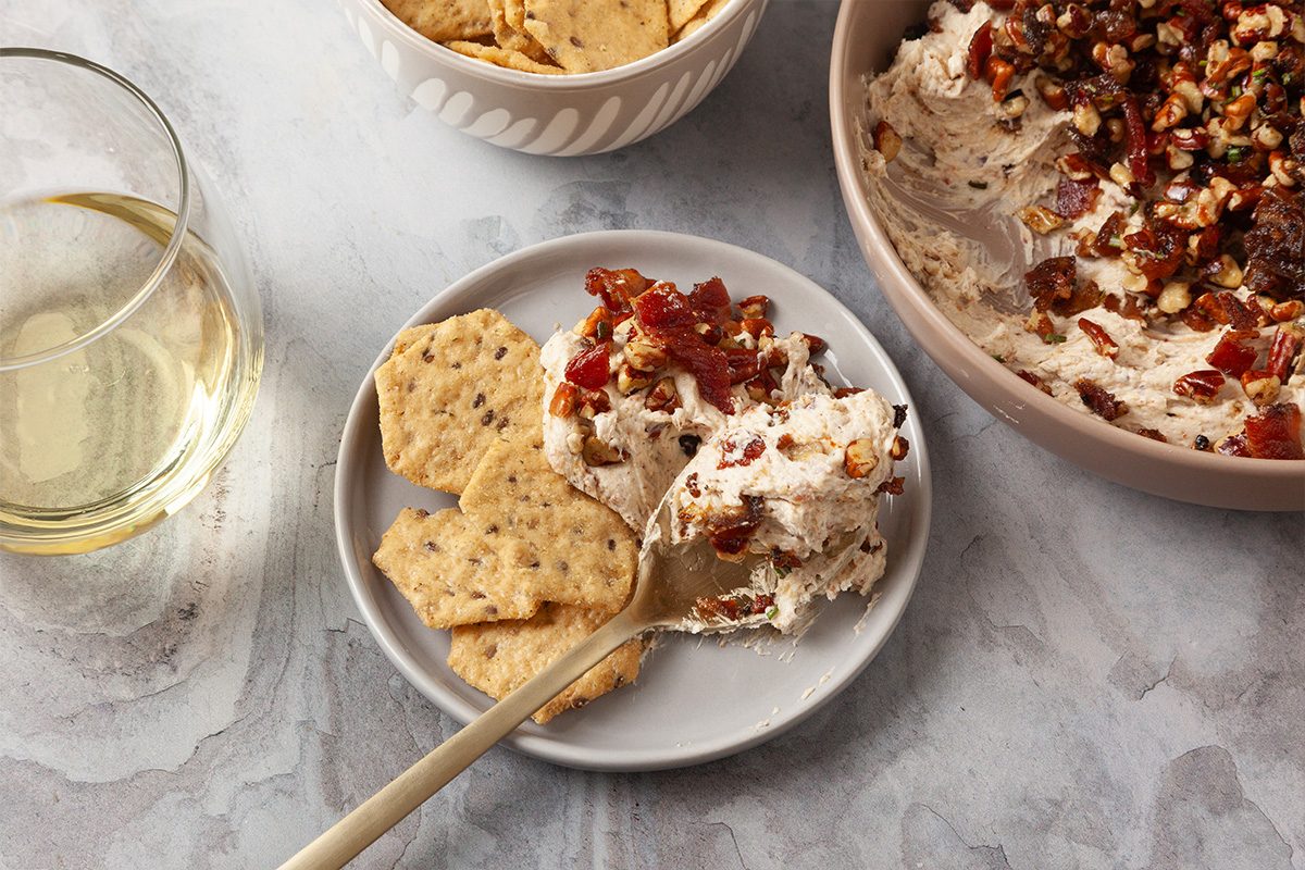 3/4th shot of a small plate with three seeded crackers topped with creamy cheese studded with nuts and dried fruit sits near a fork and white wine plus the Creamy Goat Cheese Bacon and Date Dip is seen in the background
