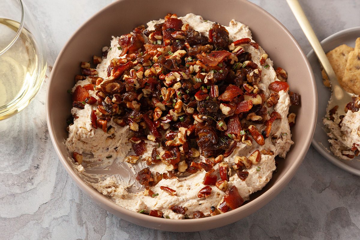 Close-up shot of Creamy Goat Cheese Bacon and Date Dip sits in a bowl with bacon nuts and herbs there is wine nearby and a small bowl with dip
