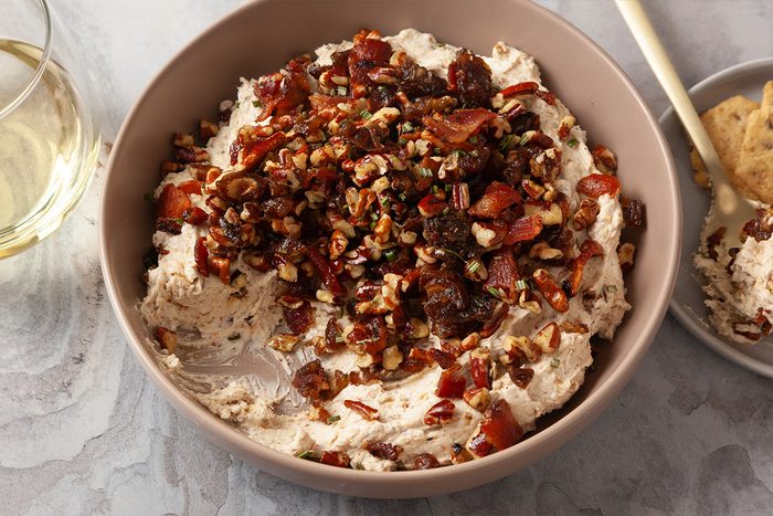 Close-up shot of Creamy Goat Cheese Bacon and Date Dip sits in a bowl with bacon nuts and herbs there is wine nearby and a small bowl with dip