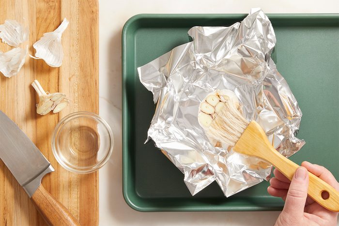 A hand brushes oil onto sliced garlic placed on aluminum foil, which sits on a baking sheet. Nearby are a chopping board, a knife, garlic cloves, and a small glass bowl.