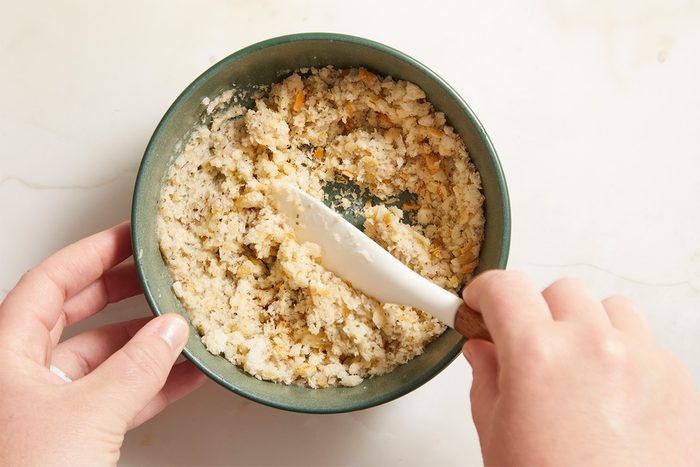 A person uses a white spatula to mix a bowl of creamy, mashed food with visible small orange and brown pieces on a light countertop.