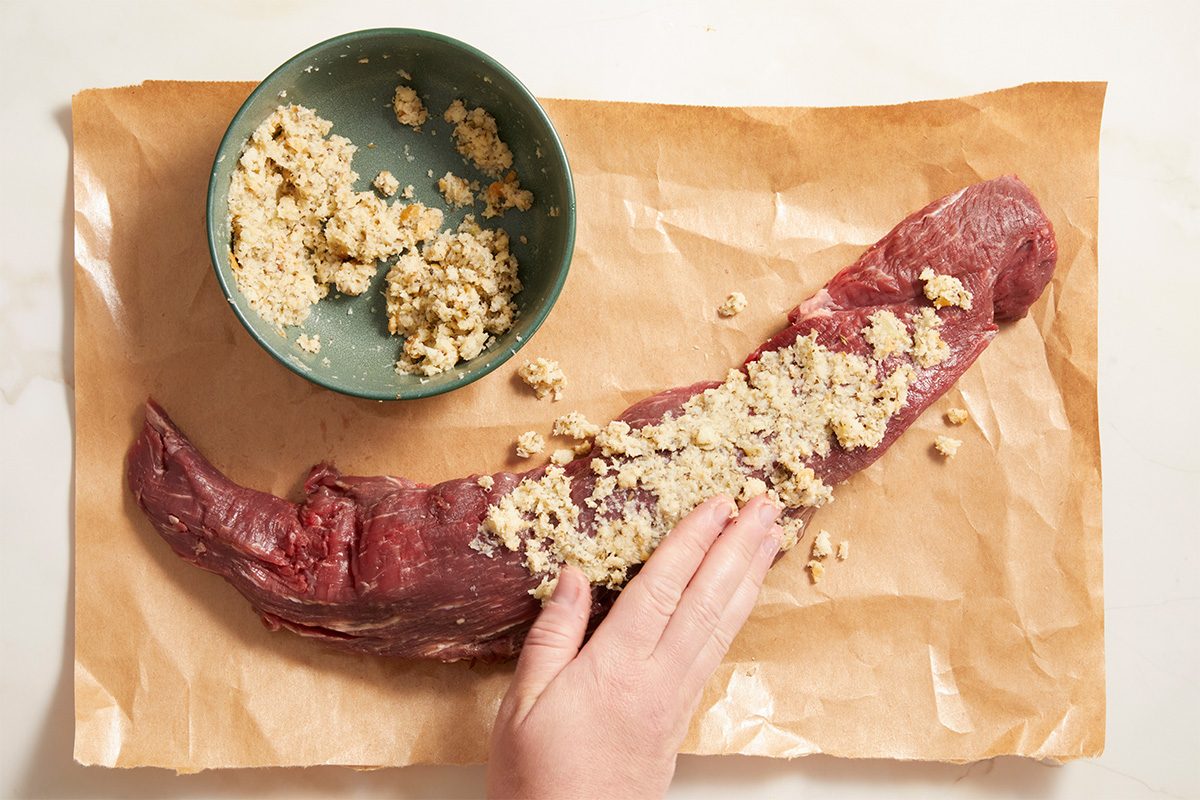 A hand spreads a breadcrumb and garlic mixture onto a raw beef tenderloin placed on brown parchment paper, with a bowl of the mixture nearby.