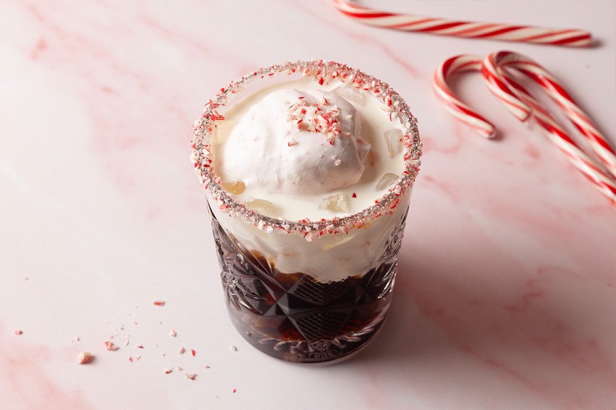 3/4th shot of a glass of Peppermint White Russian with whipped cream and crushed peppermint sits on pink marble two candy canes rest nearby