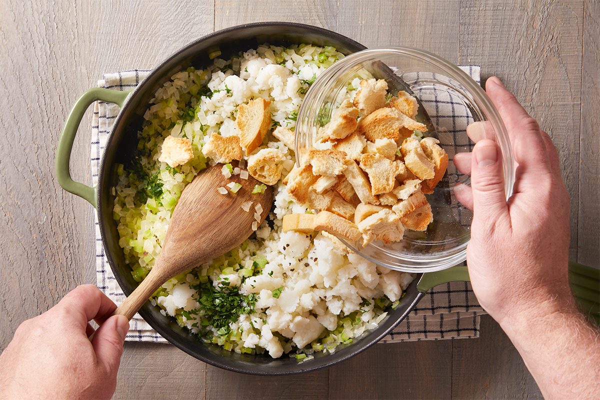 Overhead view of bread cubes being added to the skillet of sautéed vegetables, stirred together with a wooden spoon.