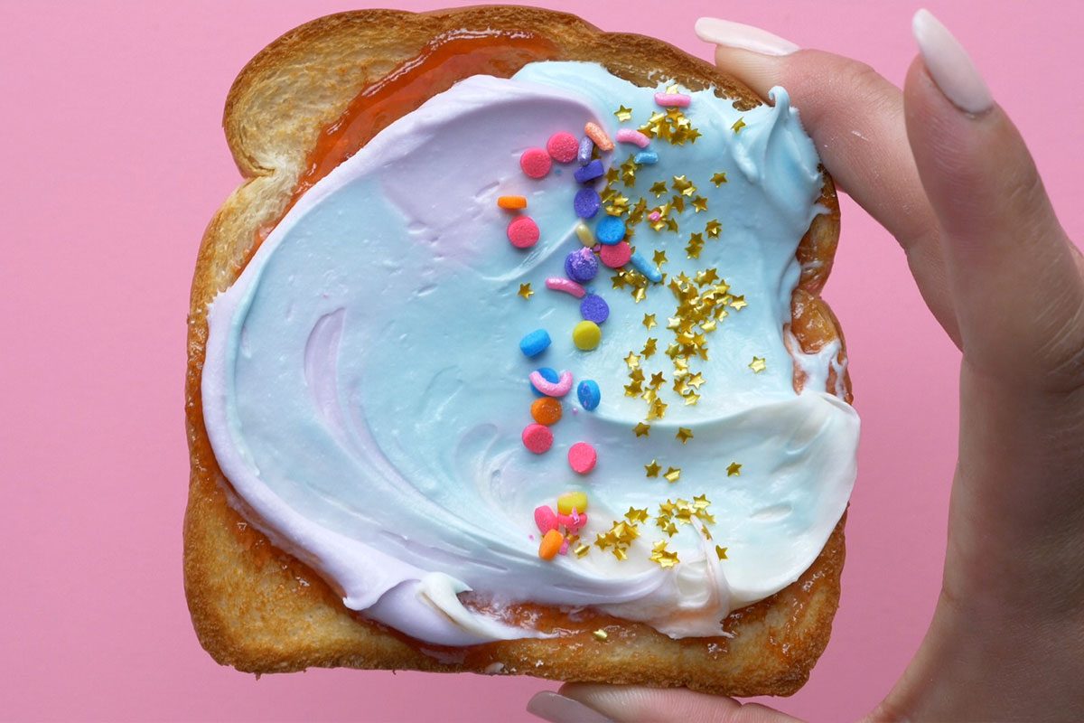 This is a close-up shot; a hand holds toast with pastel blue and white frosting; rainbow sprinkles; gold star shapes; pink background