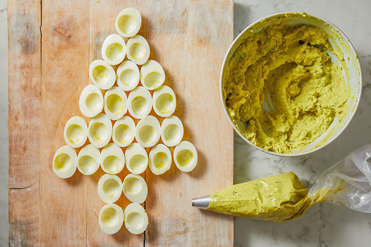 Overhead shot of a egg whites form a Christmas tree shape on a wooden board and A bowl of green filling and a piping bag sit nearby on marble