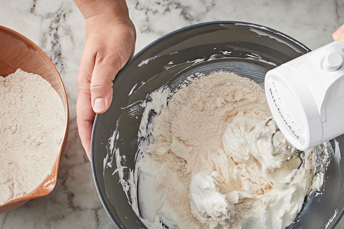 A person uses a hand mixer to blend flour into whipped egg whites in a large mixing bowl, with a bowl of more flour nearby on a marble countertop.