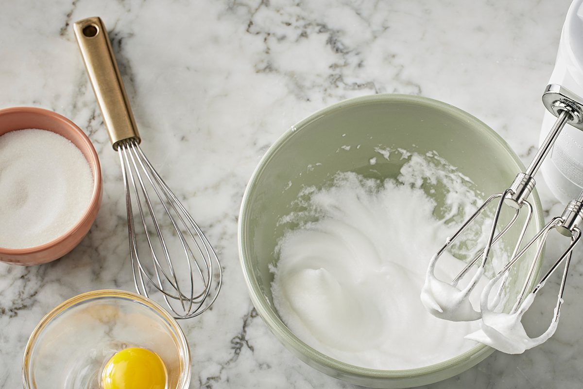 A bowl of whipped egg whites is being mixed with a hand mixer. Nearby are a whisk, a bowl with a whole egg, and a small bowl with sugar, all on a white marble countertop.