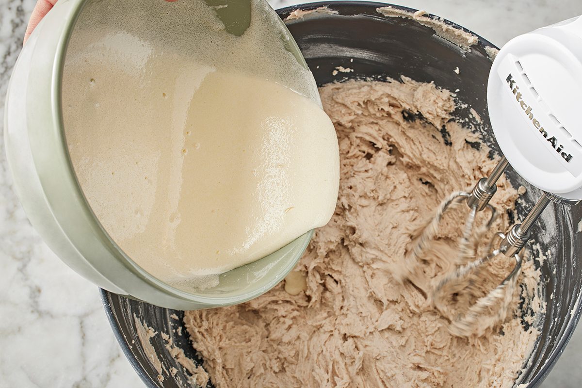 A person pours a creamy liquid from a bowl into a mixing bowl containing a thick batter, while an electric hand mixer rests nearby on the edge of the bowl.