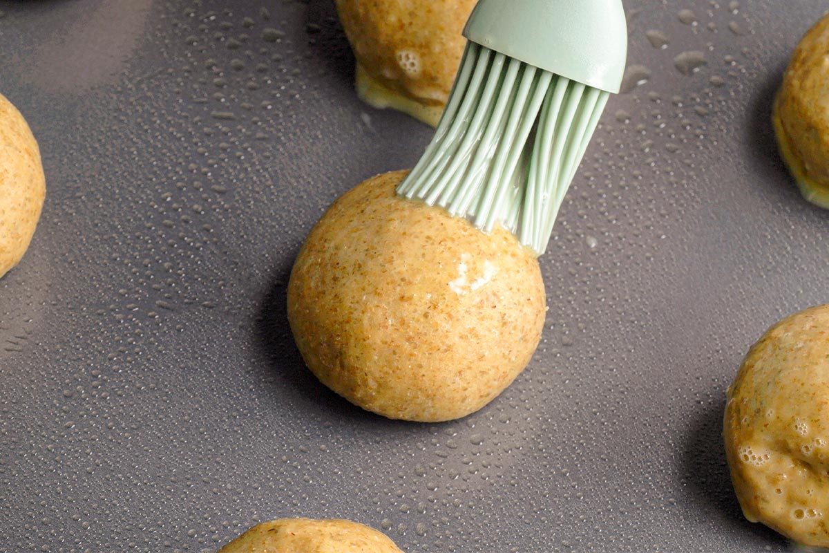 Closeup shot of a silicone brush coating a round ball of dough with egg wash on a greased baking sheet, surrounded by other unbaked dough balls;