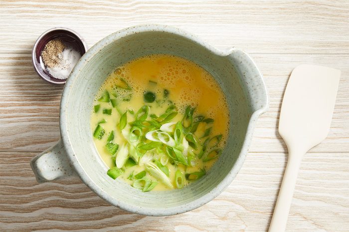 Overhead shot of whisked eggs with chopped green onions in a ceramic bowl, placed on a light wooden table with a spatula nearby.