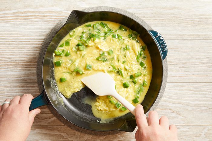 Overhead shot of eggs with green onions being stirred in a skillet, hands holding a spatula while cooking on a wooden surface.