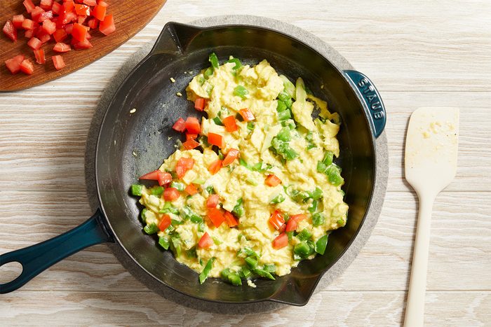 Overhead shot of eggs cooking with diced red peppers in a skillet, spatula resting beside the pan on a light wooden table.