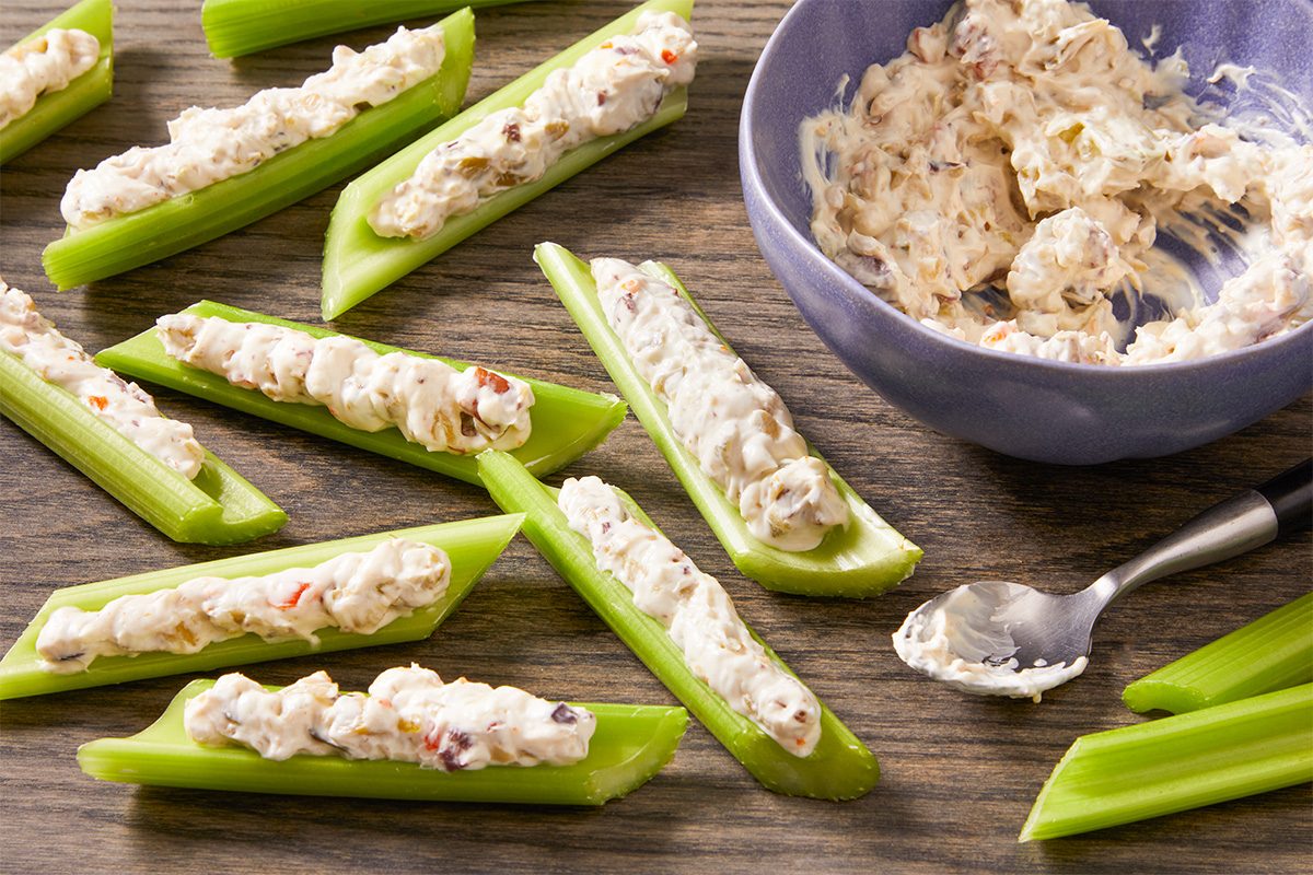 3/4 angle view shot of Olive-Stuffed Celery with crisp celery sticks filled with creamy olive-nut spread, arranged on a wooden board beside a bowl of filling and a spoon;
