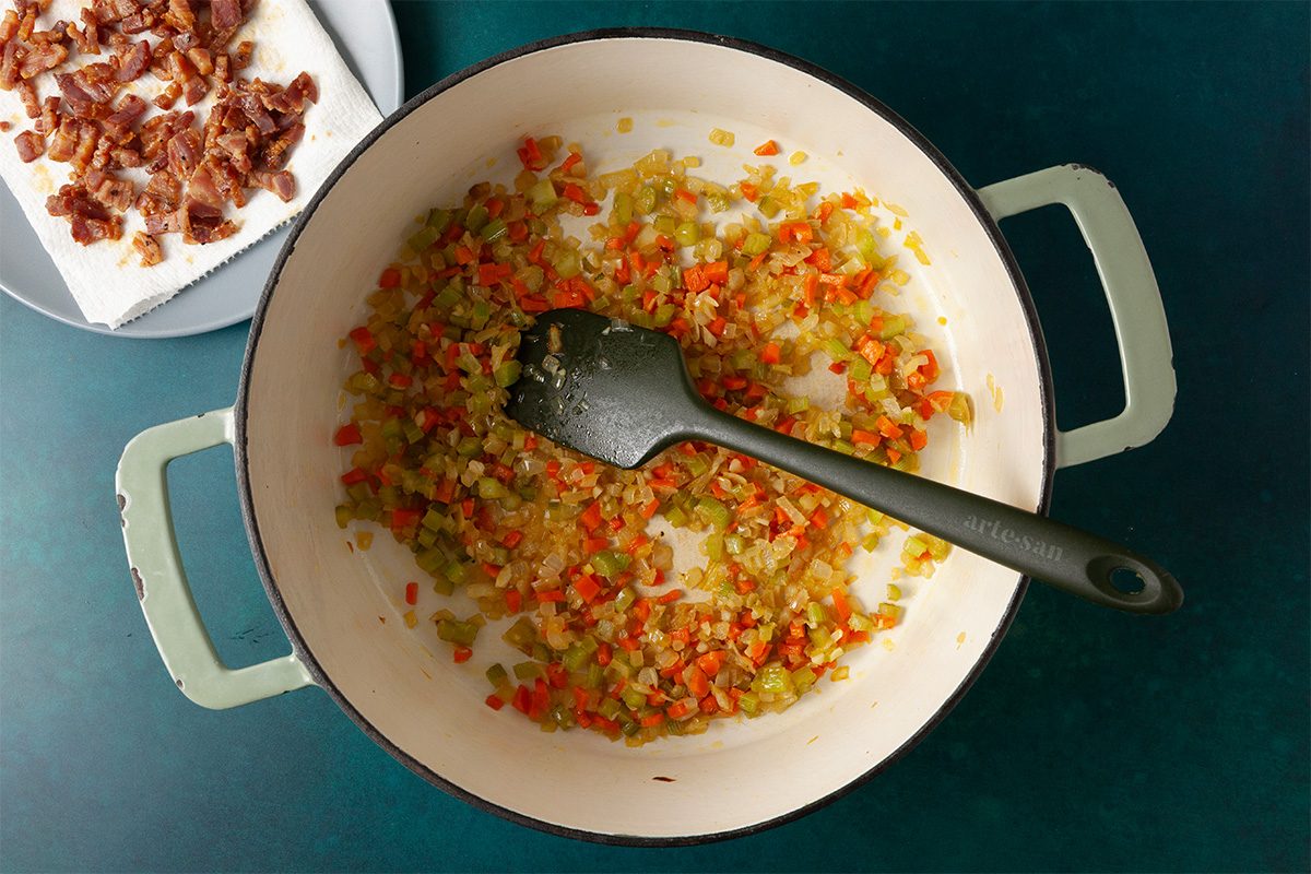 Overhead shot of a pot with sautéed chopped onions, celery, and carrots being stirred with a black spatula, while cooked bacon pieces rest on a paper towel–lined plate beside it;