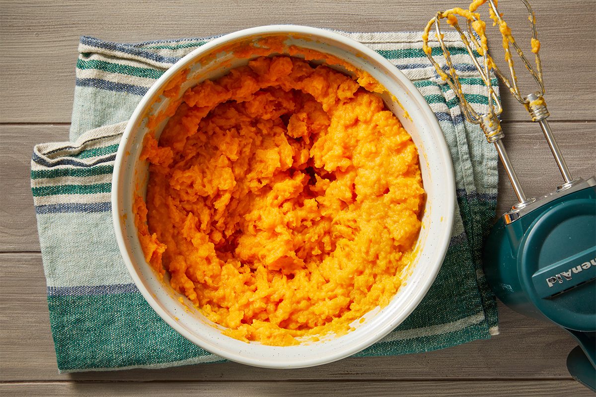 Overhead shot of a bowl of mashed sweet potatoes on a striped kitchen towel, with an electric hand mixer resting beside it on a wooden surface;