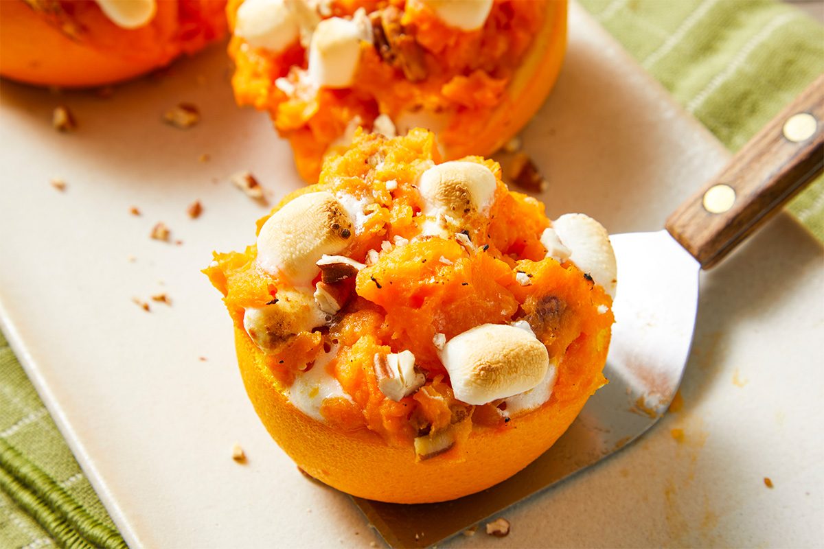 Close-up shot of a Sweet Potato Orange Cup on a serving spatula, showing the toasted marshmallows, chopped nuts, and creamy sweet potato filling inside the hollowed orange half;