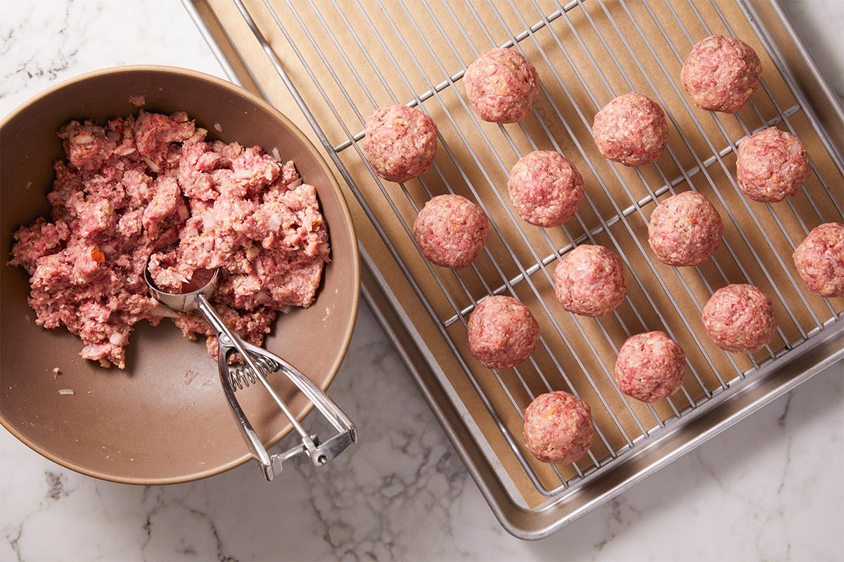 Overhead shot of a mixing bowl with raw meatball mixture and a cookie scoop sits next to a baking sheet on a marble countertop several shaped meatballs rest on the rack waiting to be cooked everything is arranged neatly for easy preparation