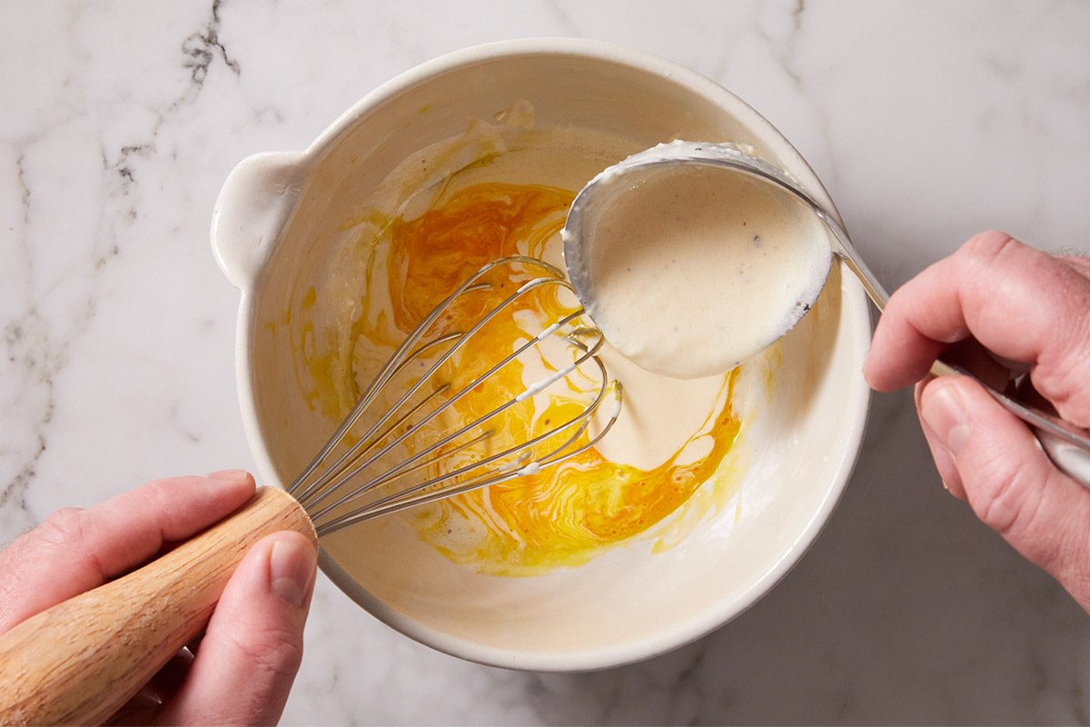 Overhead shot of a person is whisking egg yolks in a white bowl while pouring a creamy mixture from a ladle the countertop is white marble and clean