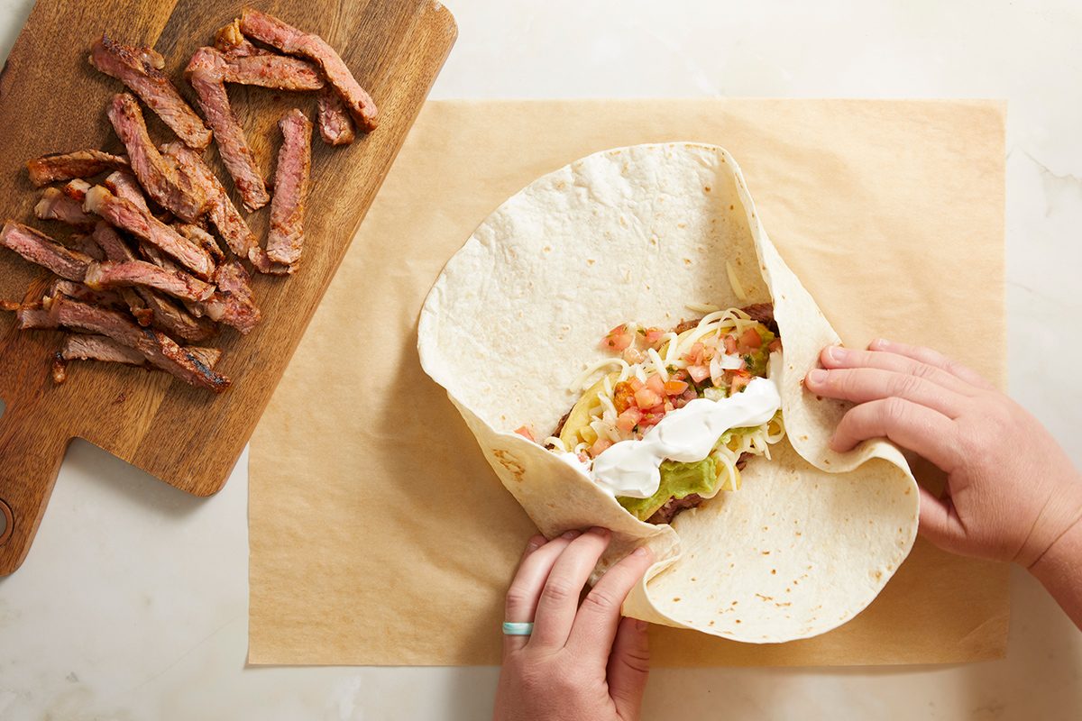 Hands folding a large flour tortilla filled with steak strips, shredded cheese, guacamole, pico de gallo, and sour cream; extra steak slices are on a wooden cutting board nearby.