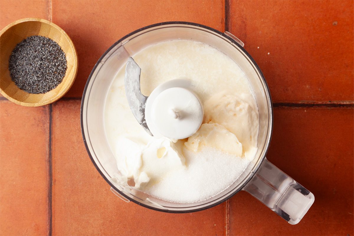 Overhead shot of a food processor blending a creamy salad dressing, with poppy seeds in a small bowl nearby on a tiled surface.