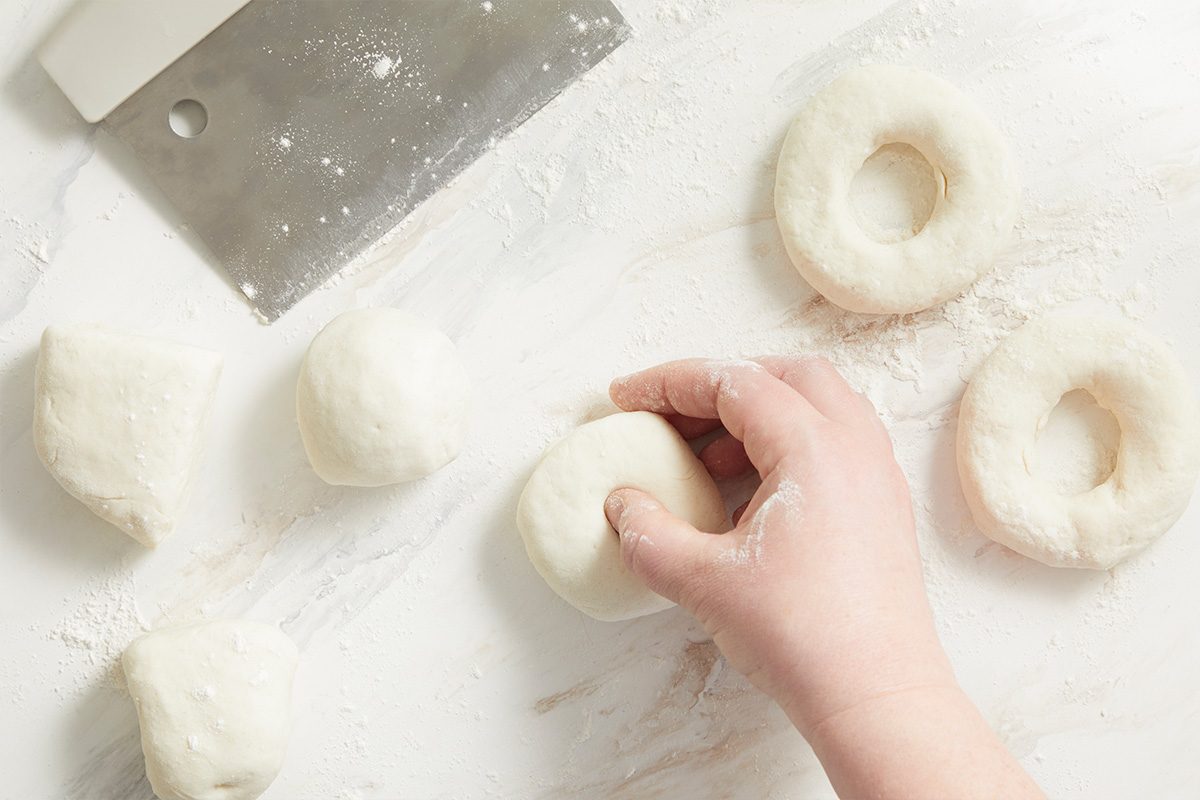 Overhead shot of a hand shaping dough into rings on a floured surface, with dough balls, a metal dough scraper, and scattered flour nearby;p