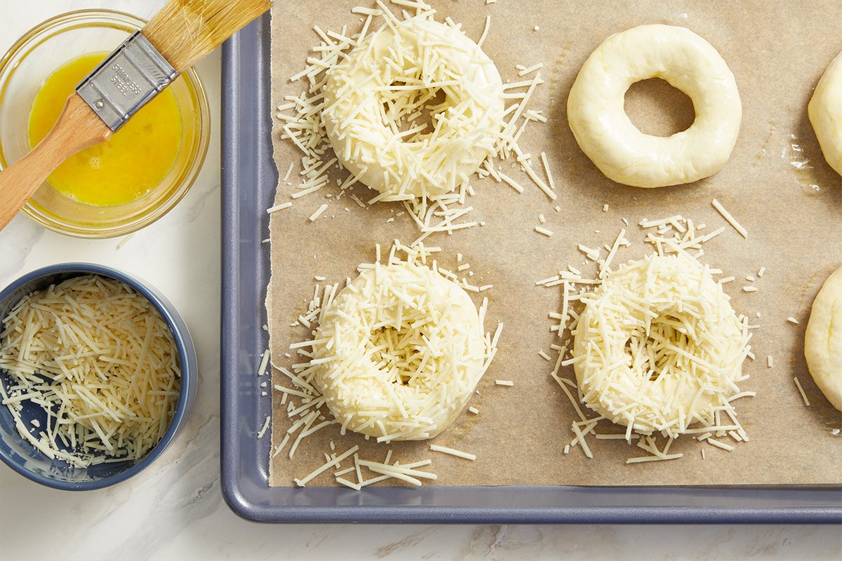 Overhead shot of a parchment-lined baking sheet with unbaked dough rings topped with shredded cheese, with bowls of shredded cheese and egg wash and a pastry brush nearby;