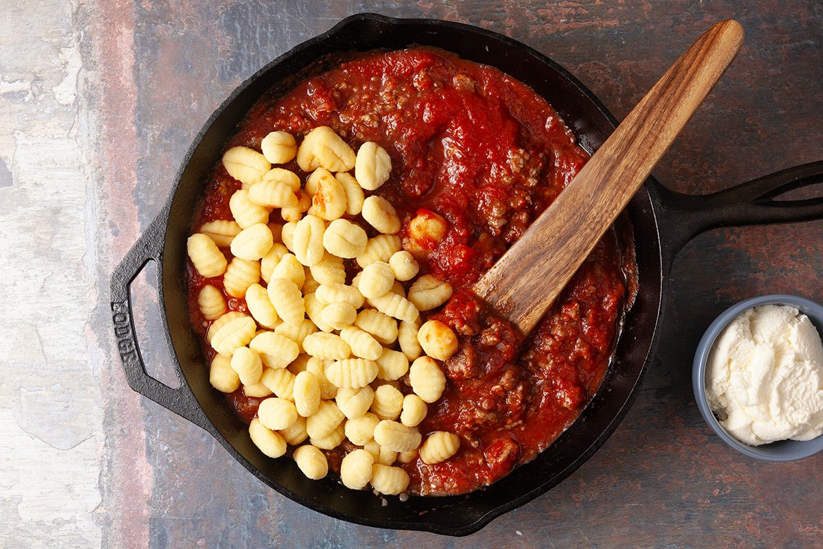 Overhead, horizontal step-by-step image of potato gnocchi added directly into a skillet of simmering sausage tomato sauce, showing the one-pan method for making gnocchi lasagna bake.
