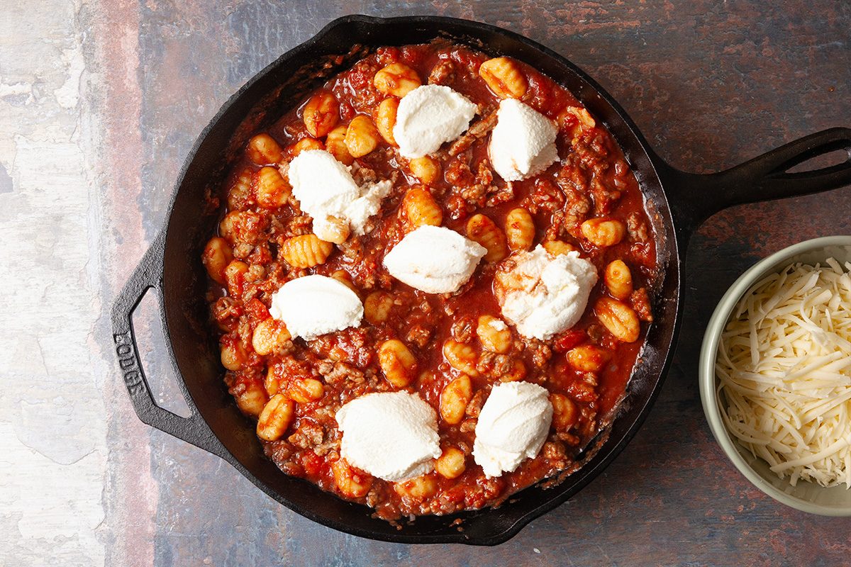 Overhead, horizontal step-by-step image of dollops of whole-milk ricotta cheese added to a gnocchi and sausage tomato mixture in a cast iron skillet before baking.