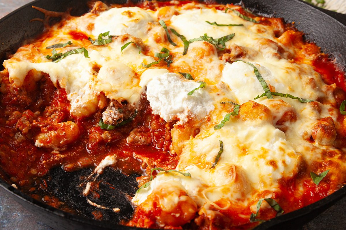 Overhead, horizontal close-up image of gnocchi lasagna bake with layers of sausage, tomato sauce, gnocchi, and melted cheese, showing texture and richness of the finished dish.