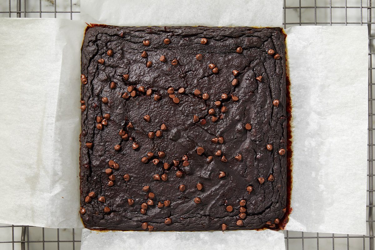 Overhead shot of baked hidden veggie brownies in a parchment-lined square pan, topped with chocolate chips and set on a cooling rack;
