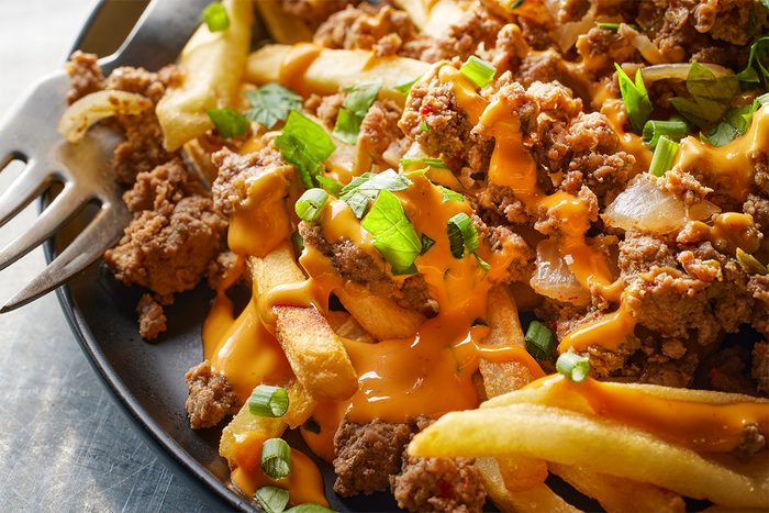 A close-up of loaded fries topped with seasoned ground beef, melted cheese sauce, and chopped green onions and cilantro on a dark plate with a fork on the side.
