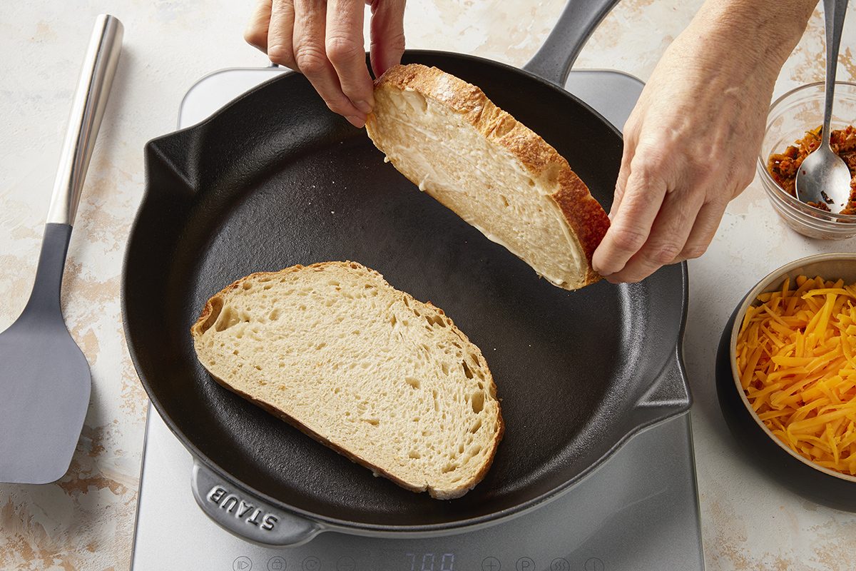 Two hands place a slice of bread into a black skillet next to another slice already in the pan; cheese and other ingredients are in small bowls nearby.