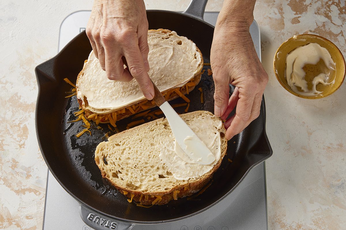 A person spreads butter on a slice of bread while assembling a sandwich with shredded cheese in a skillet on a stovetop. A small bowl with more butter sits nearby.
