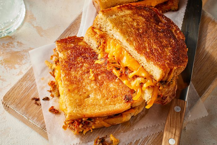 Close-up of a grilled cheese sandwich with crispy golden bread and melted cheese, cut in half and placed on parchment paper atop a wooden board with a knife beside it.