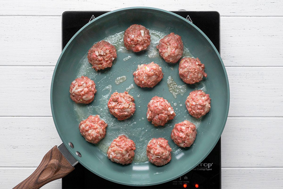 overhead shot of Raw meatballs are arranged in a single layer in a light blue frying pan, which is placed on an electric stovetop, The pan contains a small amount of oil, and the background is a white wooden surface